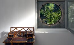 Interior view featuring a decorative wooden bench beside a round window that frames a lush green outdoor scene, highlighting natural light and contemporary design elements.