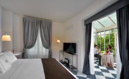 Modern hotel room featuring a comfortable bed, sleek decor, and a view of a glass-enclosed patio where a couple enjoys a meal. Ideal for relaxation and leisure.