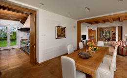 Modern dining area featuring a wooden table with a fruit centerpiece, surrounded by white chairs. The open layout connects to a kitchen and a cozy living room, showcasing natural light and elegant decor.