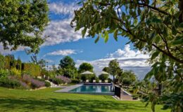 A serene outdoor landscape featuring a swimming pool surrounded by lush greenery, trees, and flowering plants under a partly cloudy sky. Comfortable lounge chairs and umbrellas are positioned near the pool, creating a relaxing atmosphere ideal for leisure and recreation.