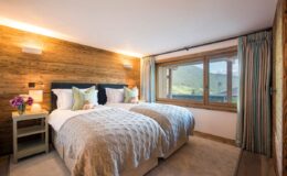 Cozy and stylish bedroom featuring two twin beds with soft linens, wooden accent walls, and a large window offering scenic views. The room is illuminated with warm lighting and decorated with tasteful accents, creating a welcoming atmosphere ideal for relaxation.
