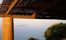 View of a serene coastal landscape featuring distant mountains and a natural wooden structure providing shade, capturing the tranquility of nature at sunset.