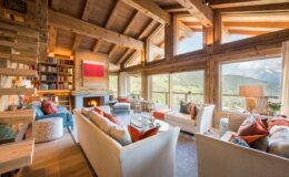 Modern alpine living room with large windows showcasing mountain views, featuring cozy seating, a fireplace, and a bookshelf. Decorated with colorful cushions, wood accents, and natural light for a warm ambiance. Ideal for luxury mountain retreats.