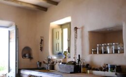 Rustic kitchen interior featuring natural clay walls, wooden beams, and a window with green shutters. The countertop showcases various cooking utensils, jars, and plants, creating a warm and inviting atmosphere.