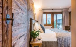 Cozy modern bedroom featuring rustic wooden walls, a comfortable bed with light bedding, and large windows allowing natural light, complemented by stylish decor and a small potted plant on a bedside table.
