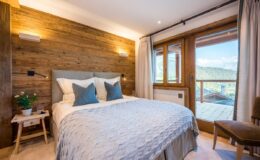 Cozy bedroom featuring a wooden accent wall, a comfortable bed with decorative pillows, and large windows that offer scenic views. The room includes soft lighting, a small bedside table with a plant, and elegant curtains, creating a warm and inviting atmosphere.