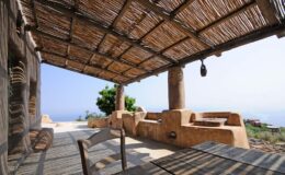 Rustic outdoor patio with a thatched roof, wooden table, and stone seating, overlooking a scenic view of the sea and landscape.