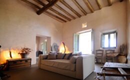 Cozy living room interior featuring a beige sofa, wooden beam ceiling, natural light from windows, and rustic decor elements.