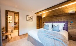 Modern bedroom interior featuring a cozy bed with decorative pillows, warm wooden accents, and soft lighting. The room includes a doorway leading to a stylish bathroom, creating a serene and inviting atmosphere. Ideal for relaxation and comfort in a contemporary setting.