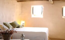 Cozy bedroom featuring a double bed with green pillows, rustic wooden beams, and natural light streaming through small windows, creating a warm and inviting atmosphere.