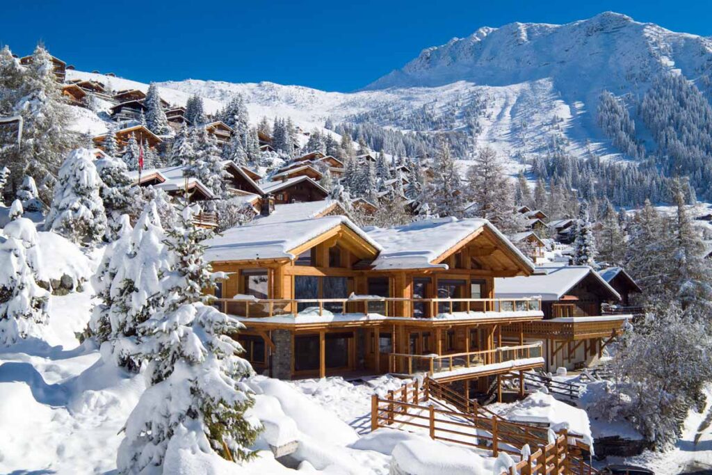 Snow-covered wooden chalet nestled in a mountainous landscape, surrounded by evergreen trees and a clear blue sky, ideal for winter vacations and outdoor activities. | Only on SopranoVillas