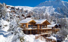 Snow-covered wooden chalet nestled in a mountainous landscape, surrounded by evergreen trees and a clear blue sky, ideal for winter vacations and outdoor activities.