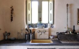 Rustic kitchen interior featuring a stone sink, wooden cabinets, and natural light streaming through open windows, creating a warm and inviting atmosphere.