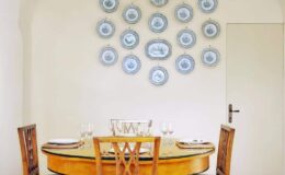 Elegant dining room featuring a round wooden table set with glassware, surrounded by four chairs. The wall is adorned with a decorative arrangement of blue and white plates, adding a touch of sophistication to the space. A closed door is visible in the background, enhancing the room's cozy atmosphere.