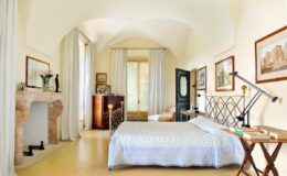 Bright and airy bedroom featuring a cozy bed with a patterned duvet, elegant decor, and large windows allowing natural light. The room includes a stylish fireplace, artwork on the walls, and a comfortable seating area, creating a serene and inviting atmosphere.