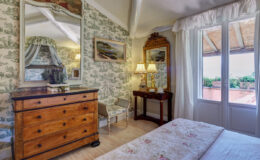 Cozy bedroom featuring vintage decor, floral wallpaper, a wooden dresser, a mirror, and large windows with scenic views. The room is elegantly lit by bedside lamps, creating a warm and inviting atmosphere.