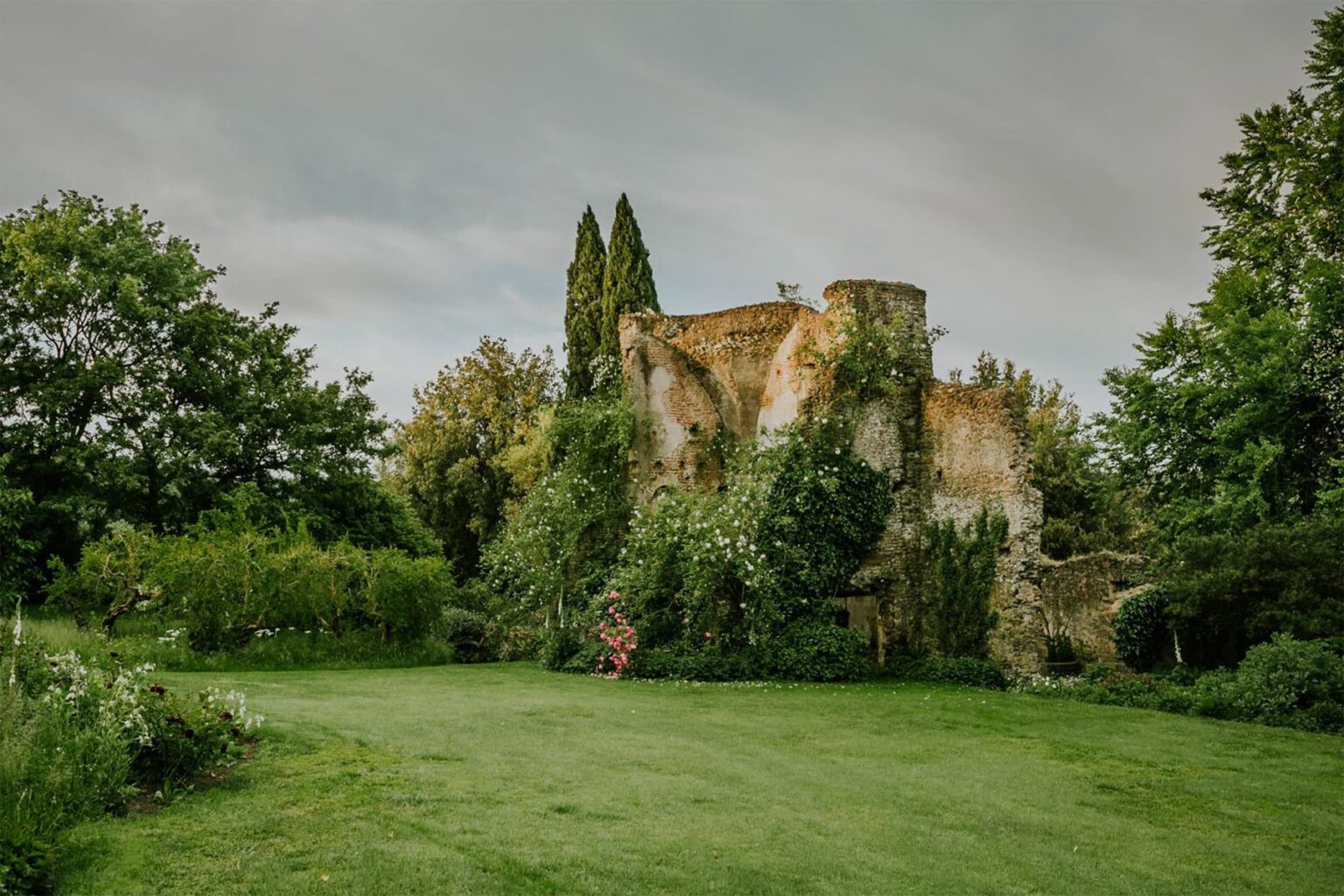 Ruins of an ancient stone structure covered in lush greenery, surrounded by trees and a manicured lawn under a cloudy sky. | Only on SopranoVillas