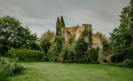 Ruins of an ancient stone structure covered in lush greenery, surrounded by trees and a manicured lawn under a cloudy sky.