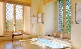 Bright and airy bathroom featuring large diamond-patterned windows, a soaking tub, a wooden chair, and neatly hung white towels. The warm color palette creates a serene atmosphere, perfect for relaxation and rejuvenation.