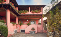 Charming pink villa with flower-adorned balconies and stone accents, set against a clear sky, showcasing a welcoming entrance and landscaped surroundings.