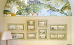Elegant wall display featuring a large mural of a serene landscape above a collection of eight framed artworks depicting various scenic views, complemented by a stylish lamp in a well-lit room.