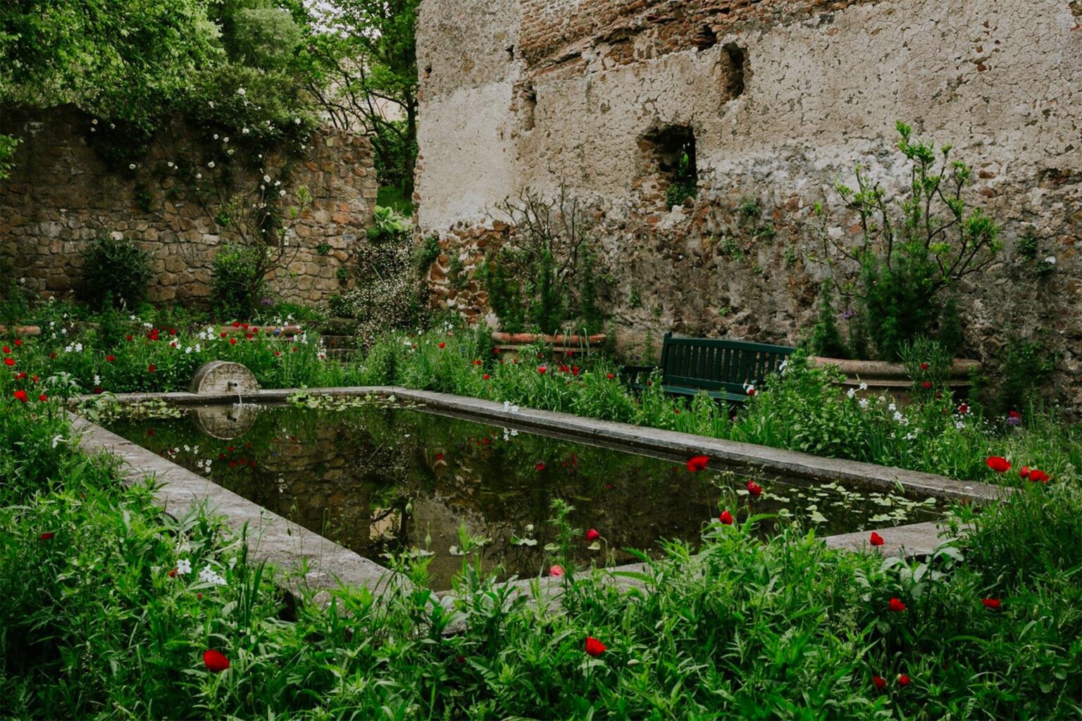 Serene garden scene featuring a small pond surrounded by lush greenery and blooming flowers, with rustic stone walls in the background. | Only on SopranoVillas