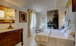Cozy bedroom featuring a white bed with decorative pillows, a wooden dresser, a vintage lamp, and large windows with elegant curtains. A landscape painting adorns the wall, adding a touch of sophistication to the inviting atmosphere.