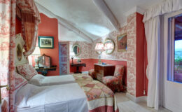 Cozy guest room featuring two twin beds, vintage decor, and elegant wallpaper in warm tones, complemented by a writing desk and scenic views through a large window. Ideal for relaxation and comfort.