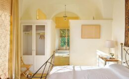 Bright and airy bedroom featuring a white bed, light-colored walls, and a cozy atmosphere. The room includes a wardrobe, a small table with a lamp, and a view of a green outdoor area through a window. Ideal for relaxation and tranquility.