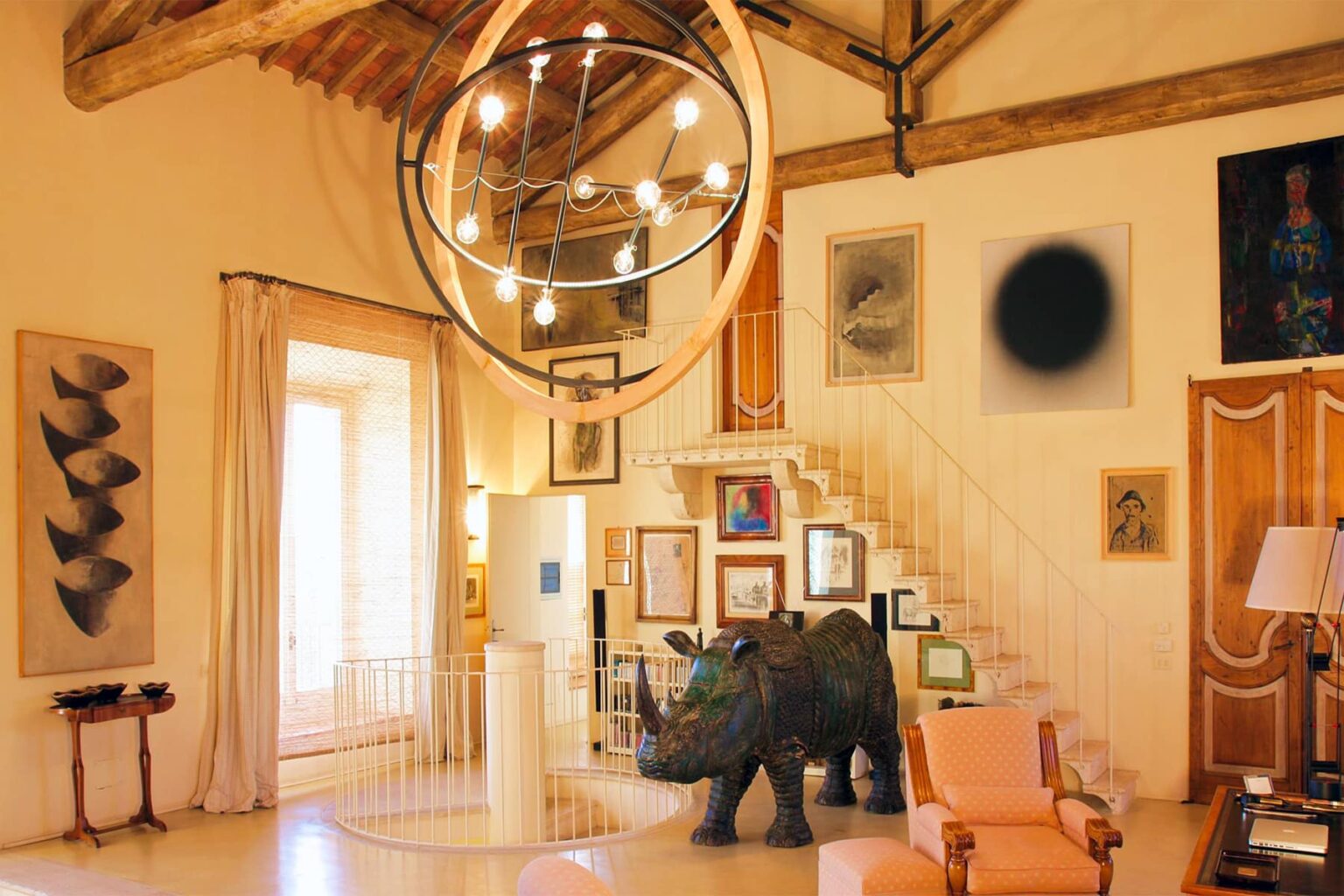 Modern living room with artistic decor featuring a large circular chandelier, various framed artworks on the walls, a decorative elephant sculpture, and stylish furniture. Bright natural light filters through large windows, enhancing the inviting atmosphere. | Only on SopranoVillas