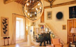 Modern living room with artistic decor featuring a large circular chandelier, various framed artworks on the walls, a decorative elephant sculpture, and stylish furniture. Bright natural light filters through large windows, enhancing the inviting atmosphere.
