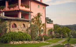 Charming pink villa with stone accents surrounded by lush greenery and a well-maintained garden, featuring a stone pathway leading to the entrance, set against a scenic hillside backdrop.