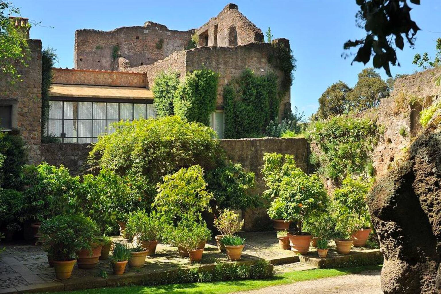 Lush garden with potted plants surrounded by ancient stone walls and ruins under a clear blue sky. | Only on SopranoVillas