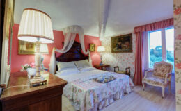 Cozy bedroom featuring a floral bedspread, elegant lamp, and vintage decor with pink walls and large windows allowing natural light. Ideal for a relaxing getaway.