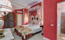 Cozy hotel room featuring twin beds with decorative bedding, vibrant red walls, elegant curtains, and antique furnishings. The space includes a bedside lamp and a stylish wardrobe, creating a warm and inviting atmosphere.