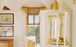 Bright and airy interior of a stylish room featuring white walls, large windows with yellow drapes, a wooden dresser, and framed artwork. A glass display cabinet showcases decorative items, creating an inviting atmosphere.