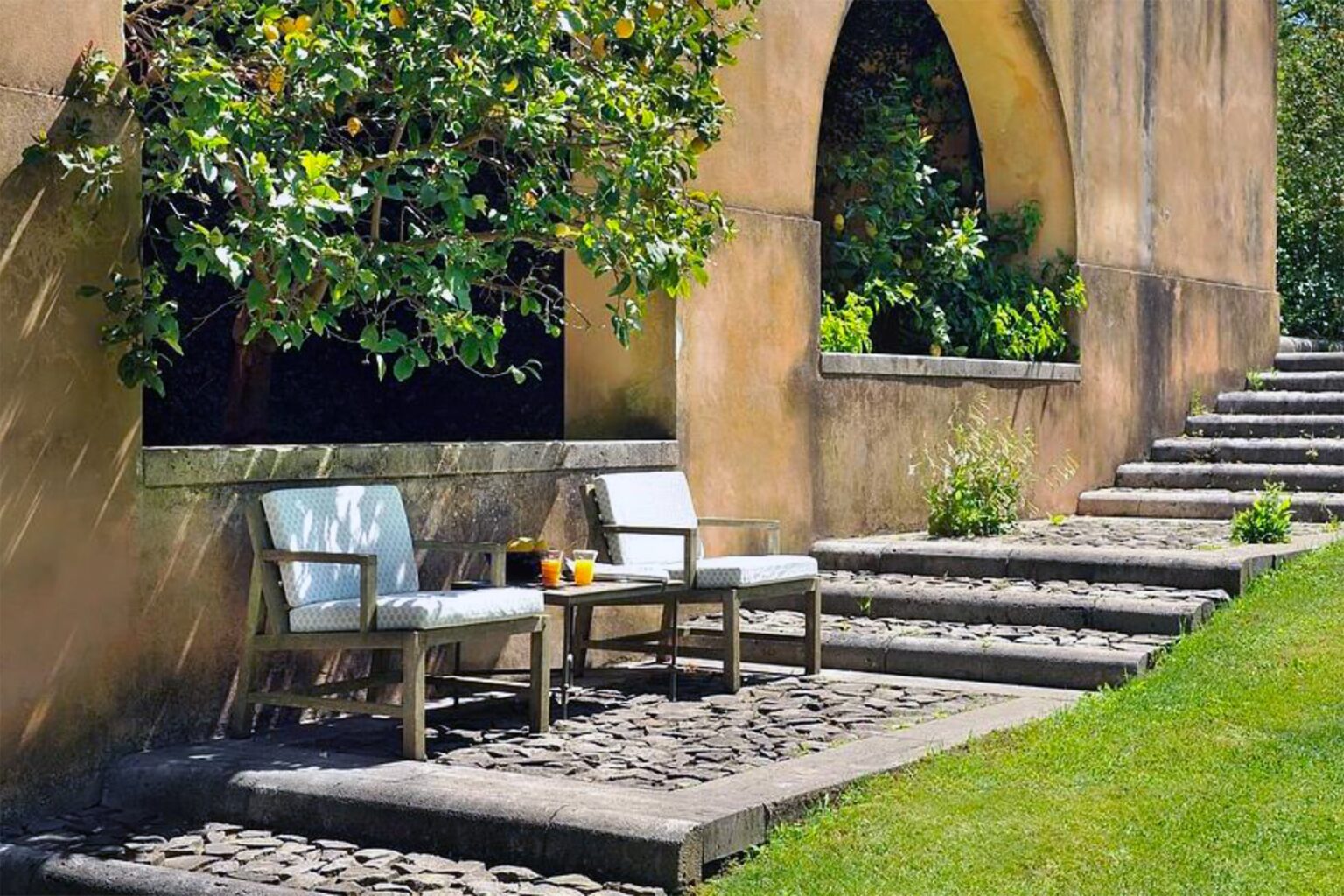 Outdoor seating area with two white chairs beside a stone pathway, surrounded by lush greenery and a textured wall. Ideal for relaxation in a serene garden setting. | Only on SopranoVillas