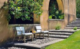 Outdoor seating area with two white chairs beside a stone pathway, surrounded by lush greenery and a textured wall. Ideal for relaxation in a serene garden setting.