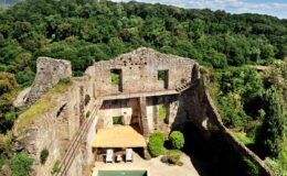 Ruins of an ancient stone castle surrounded by lush green forest, featuring an outdoor seating area and a swimming pool in the foreground.