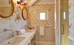 Elegant bathroom featuring vintage decor with floral wallpaper, dual sinks, and ornate mirrors. The space includes a glass shower and stylish lighting, creating a cozy and inviting atmosphere.