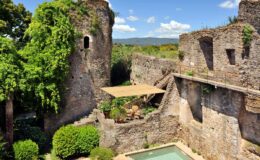 Ruins of an ancient stone castle surrounded by lush greenery and a clear blue sky, featuring a small pool and a shaded area, showcasing historical architecture and natural beauty.