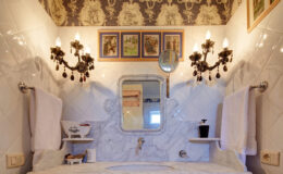 Elegant bathroom interior featuring marble countertops, vintage-style sconces, and artistic wall decor, including framed pictures and patterned wallpaper. Soft towels and a stylish mirror enhance the luxurious ambiance.