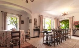 Elegant dining room featuring a large wooden table surrounded by chairs, with natural light streaming through arched windows. Decorated with floral curtains, vintage artwork, and a chandelier, the space exudes a warm and inviting atmosphere.