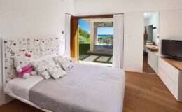 Cozy bedroom featuring a plush bed with decorative pillows, a Hello Kitty stuffed animal, and a modern TV. The room opens up to a stunning outdoor view, showcasing a patio area and lush greenery, creating a serene atmosphere.