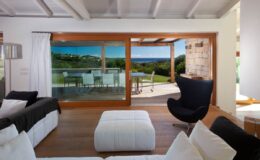 Modern living room with large windows showcasing a scenic outdoor view, featuring stylish furniture, including a black chair and white ottoman, and bright natural light enhancing the space.