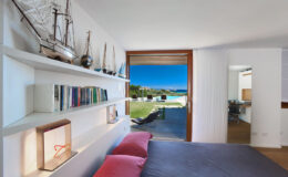 Modern bedroom with a view, featuring a cozy bed with pink pillows, a bookshelf filled with books, and decorative sailing ships. Large glass doors open to a scenic outdoor area with a lawn and ocean view, emphasizing a bright and airy atmosphere.