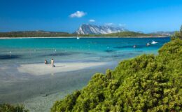 Scenic beach view featuring turquoise waters, a sandy shore, and lush greenery, with a mountain in the background and two people enjoying the shoreline. Ideal for travel and nature enthusiasts.