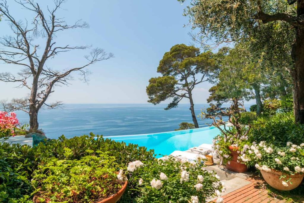 Scenic view of a tranquil infinity pool surrounded by lush greenery and colorful flowers, overlooking a serene ocean landscape under a clear blue sky. Ideal for relaxation and luxury living. | Only on SopranoVillas