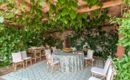 Cozy outdoor dining area adorned with lush greenery, featuring a round table with a decorative tablecloth and fruit centerpiece, surrounded by elegant chairs, perfect for gatherings or events.