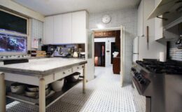 Modern kitchen interior featuring a central island with bowls, stainless steel stove, and a light color scheme. The walls are adorned with white tiles, and a clock hangs above the doorway leading to another room.